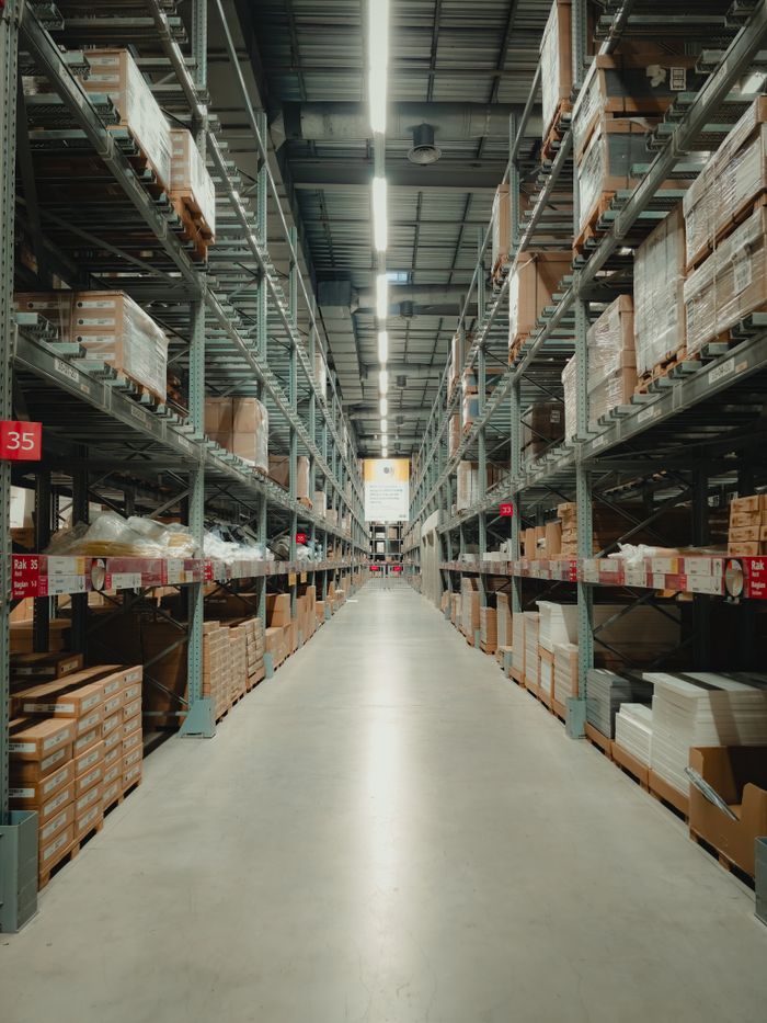 footer-cta-img Spacious warehouse aisle with shelves filled with neatly stacked products and pallets.