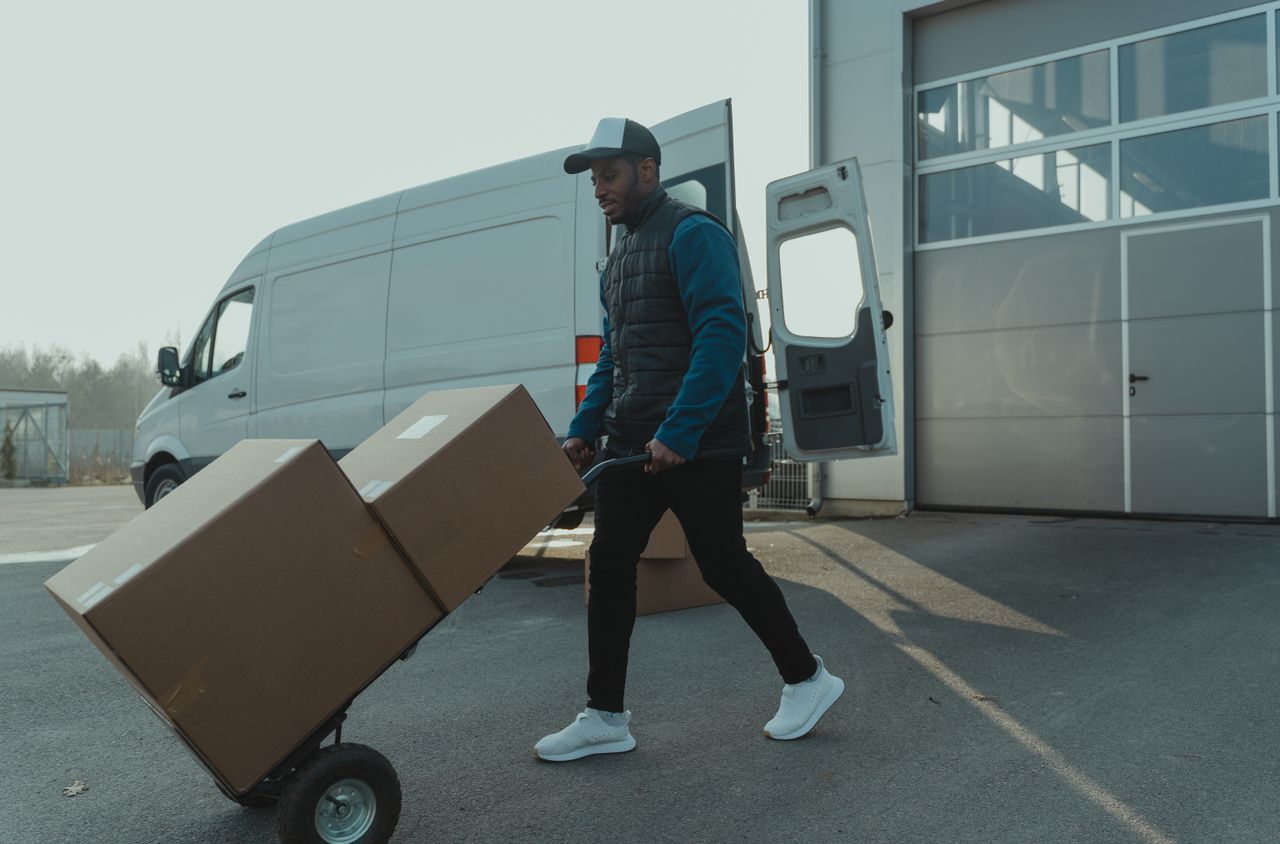 gallery-03 Courier pushing a trolley with boxes near a van at a warehouse, showcasing delivery services.